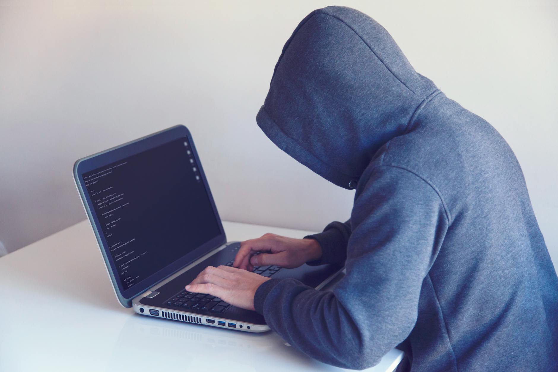 Haker w kapturze przy biurku pracuje na laptopie w jasnym pokoju